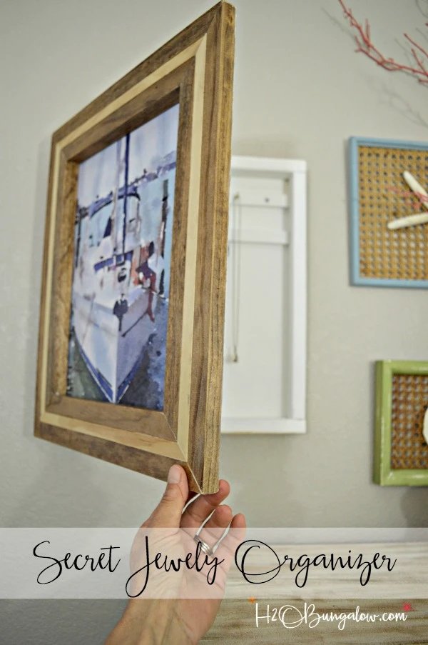 A DIY wall-mounted secret jewelry organizer featuring a decorative wood-framed mirror on hinges that opens to reveal a shallow cabinet with rows of hooks for necklaces.