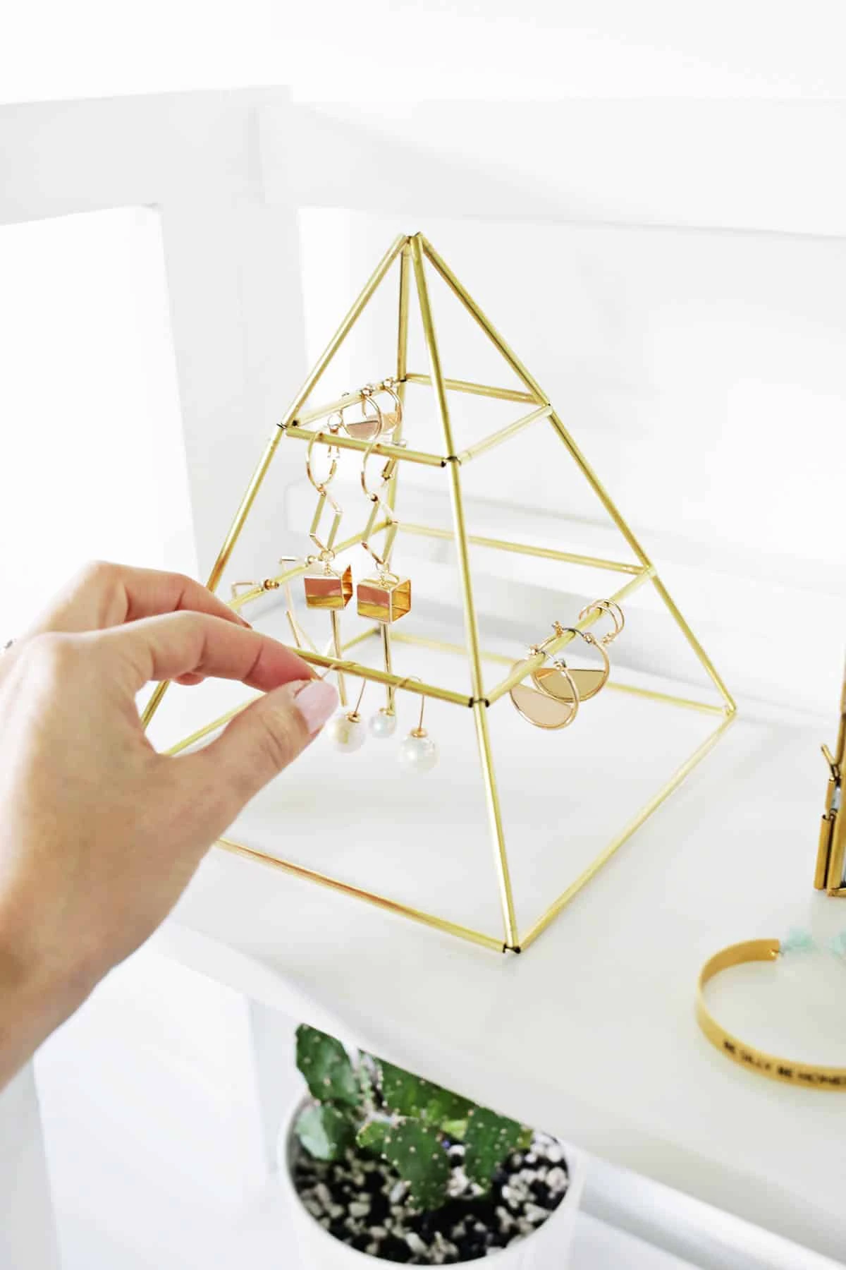 A modern geometric tabletop jewelry organizer shaped like a pyramid made of thin brass tubing, holding delicate gold earrings and bracelets.