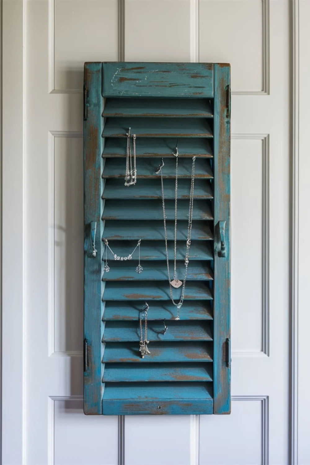 A teal repurposed wooden window shutter mounted on a wall, functioning as a DIY jewelry organizer with earrings hanging from the louvers and necklaces on hooks.