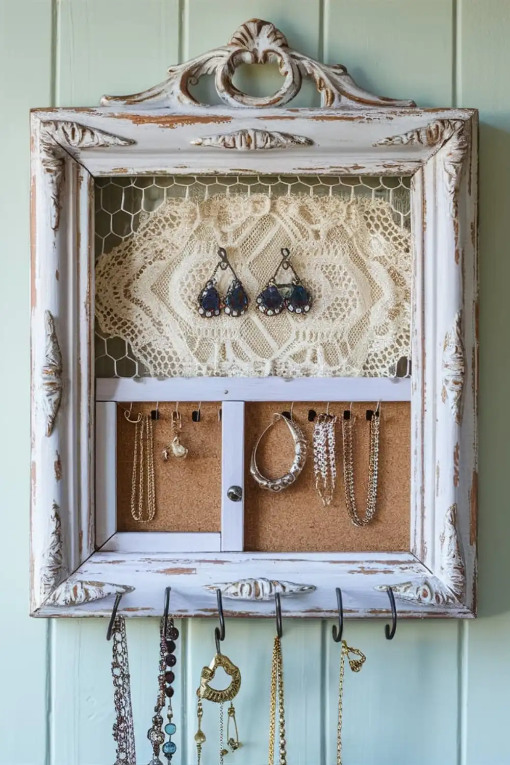 A teal repurposed wooden window shutter mounted on a wall, functioning as a DIY jewelry organizer with earrings hanging from the louvers and necklaces on hooks.