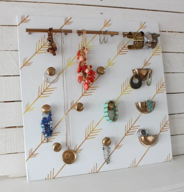A DIY wall-mounted jewelry organizer featuring an eclectic mix of gold-toned cabinet knobs and pulls mounted on a white backboard with a gold arrow pattern, displaying necklaces and bracelets.