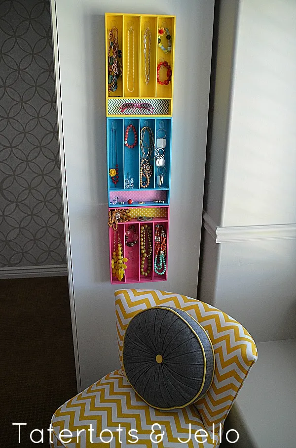 A repurposed wooden silverware tray mounted vertically on a wall as a jewelry organizer, featuring painted compartments and hooks for necklaces.