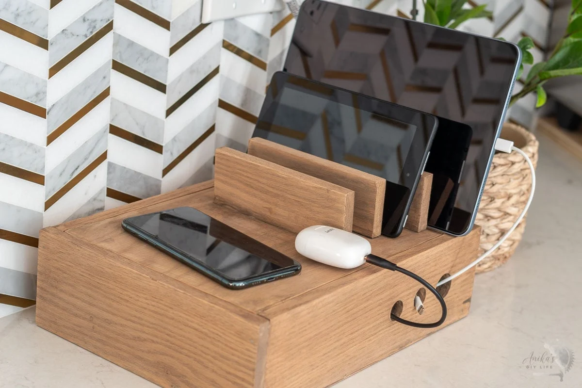 A wooden charging station box on a kitchen counter holding multiple phones and a tablet, hiding the charging cords inside.