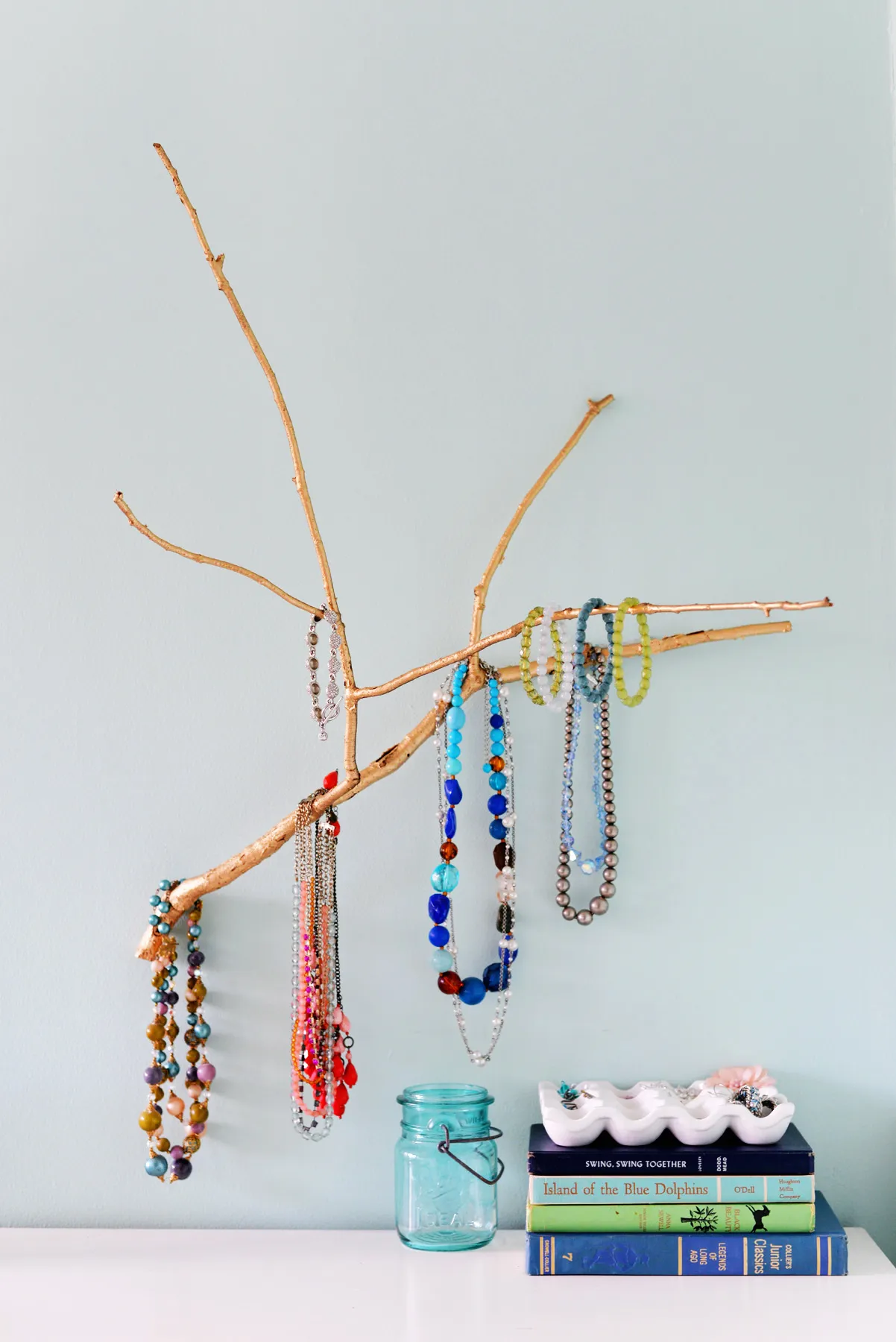 A sturdy tree branch spray-painted and mounted horizontally to a wall, functioning as a rustic DIY jewelry organizer for necklaces and bracelets.