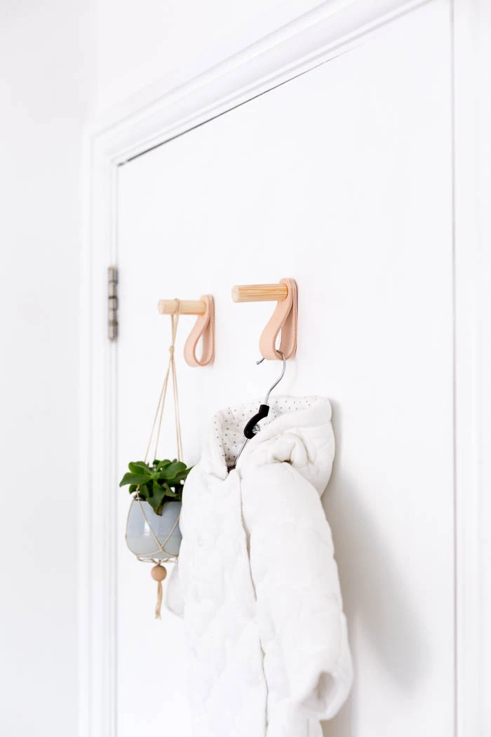 Two modern DIY wall hooks made from light wood dowels and looped tan leather straps, mounted on a white door and holding a small hanging planter and a white jacket.