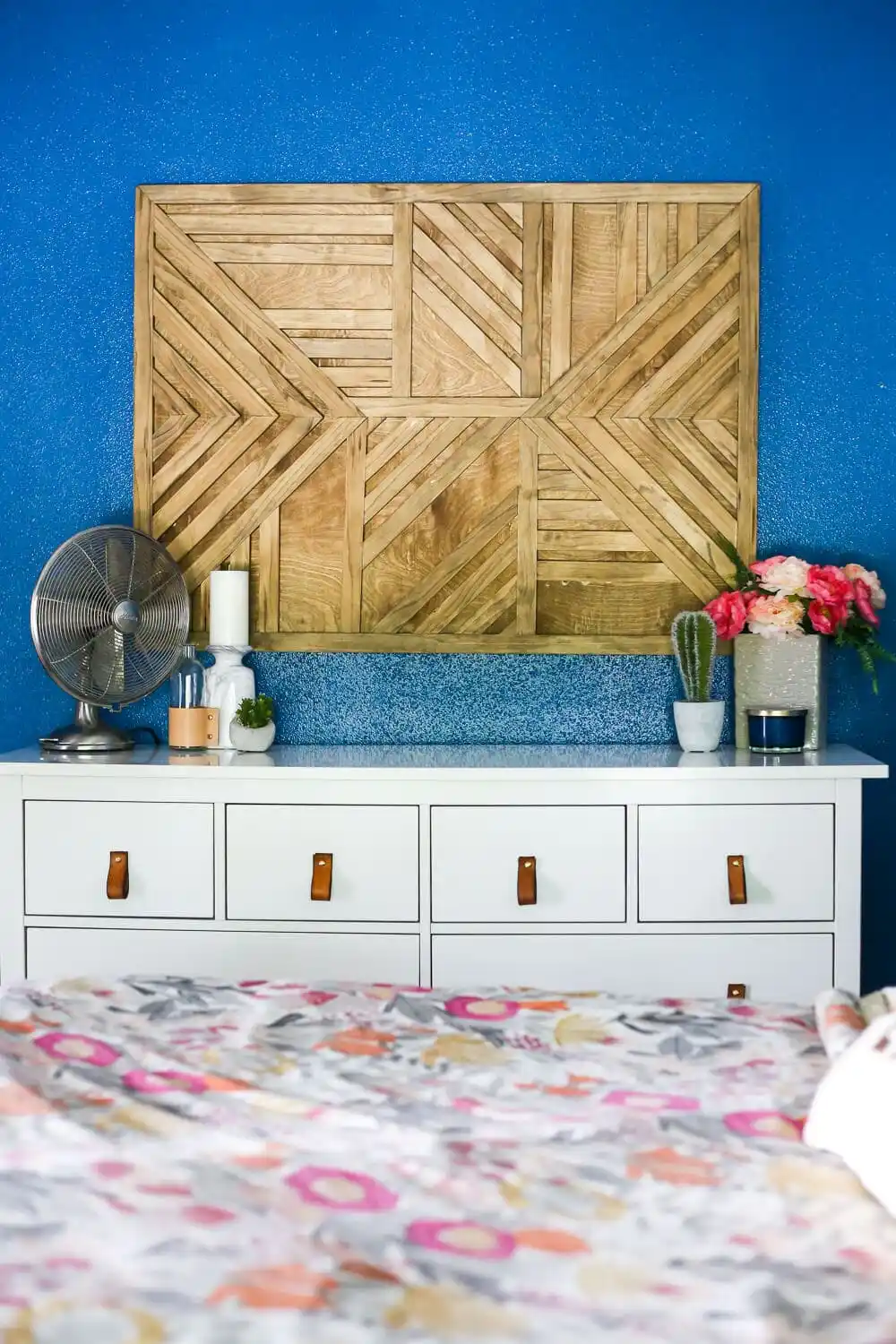 A handmade geometric wood wall art piece made of angled wood strips hanging on a bright blue wall above a white bedroom dresser, serving as a cute DIY room decor project.
