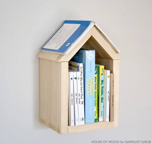 Small, minimalist house-shaped wooden shadow box shelves holding books on a light-colored bedroom wall.