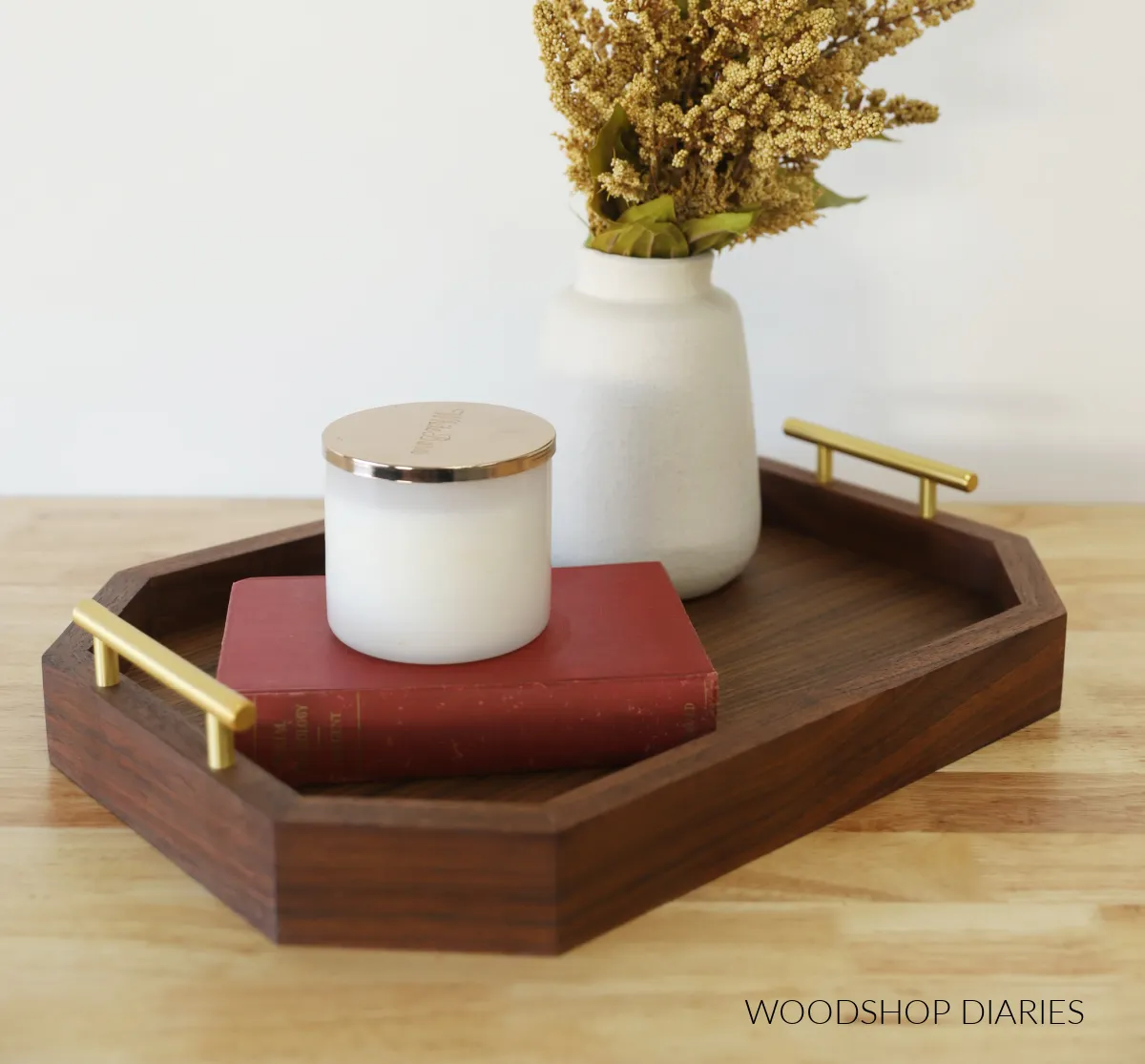 A modern, octagonal dark wood serving tray with gold handles holding a red book, a white candle, and a vase of dried flowers, making for a stylish and sweet wooden gift for home decor.
