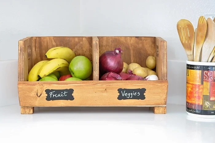 A stained wooden divided produce storage bin sitting on a white kitchen counter. The left compartment is labeled "Fruit" and holds bananas and apples, while the right compartment is labeled "Veggies" and holds red onions and potatoes.