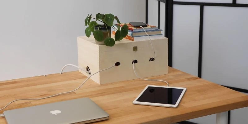 A handmade light plywood cable management box sitting on a wooden desk. The box features three arched notches on the front where charging cables emerge to power a tablet and phone. A small potted plant and books are placed on top of the box.