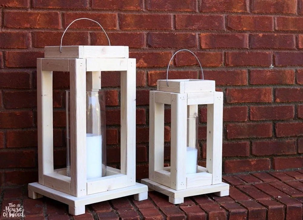 wo open-frame wooden lanterns painted white, sitting on a red brick ledge. They feature a tiered base with small feet and a tiered top cap with a wire handle. Each holds a glass hurricane vase with a white pillar candle inside.