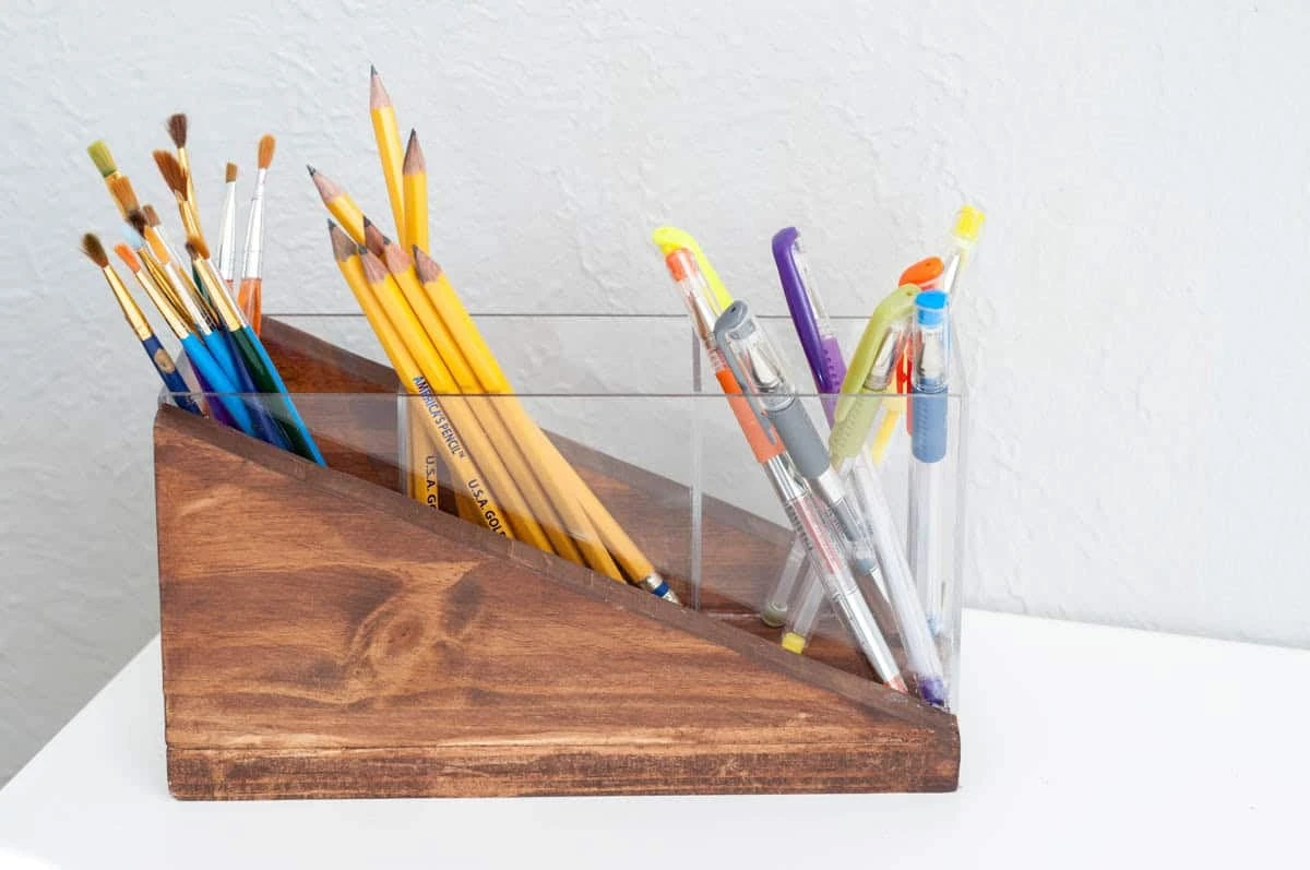 A modern, handmade pencil holder sitting on a white desk, constructed from an angled block of dark-stained wood with a clear acrylic front panel containing colored pens and paintbrushes.