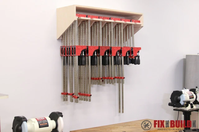 A wall-mounted wooden clamp rack built from scrap plywood, featuring angled brackets holding a neatly organized collection of large red parallel clamps above a shop workbench.
