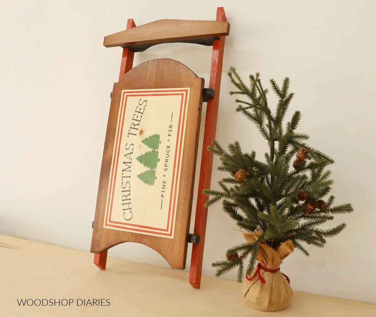 Handmade decorative wooden sled with red rails and a Christmas Trees stencil leaning next to a burlap-wrapped mini pine tree 